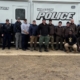 A group of 20 law enforcement officers, including two K9 officers, pose for a photo in front of the Williston Police Department's white Mobile Command Center trailer.