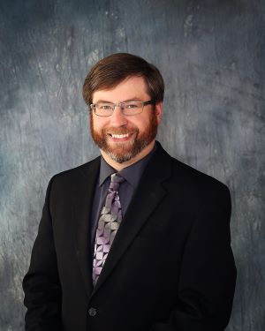 Jordan Wahl wears a black suit coat with purple tie and black wire-rimmed glasses while smiling for his professional headshot. The background is a gray blue color.