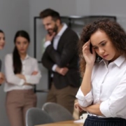 A woman standing alone wears a white shirt is in the right-hand corner. She is in the foreground. She has bent her elbow to place her fingertips to her temple while her chin is tilting down, emoting signs of stress or worry. In the left-hand corner are three people, two women and one man, who appear to be talking while looking at the woman standing alone. These people are in the background, and they are out of focus.