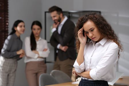 A woman standing alone wears a white shirt is in the right-hand corner. She is in the foreground. She has bent her elbow to place her fingertips to her temple while her chin is tilting down, emoting signs of stress or worry. In the left-hand corner are three people, two women and one man, who appear to be talking while looking at the woman standing alone. These people are in the background, and they are out of focus.