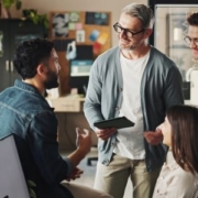 Four employees closely gather around a desk. They are smiling and appear engaged. One is sitting in an office chair adjacent to a computer monitor, one is sitting on the corner of the desk, and the remaining two are standing.