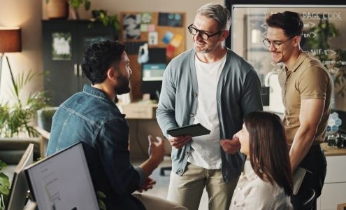 Four employees closely gather around a desk. They are smiling and appear engaged. One is sitting in an office chair adjacent to a computer monitor, one is sitting on the corner of the desk, and the remaining two are standing.